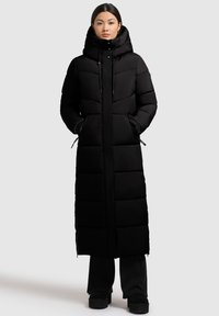 Ilgas juodas pūstas paltas su aukštu kaklu, užtrauktuku ir reguliuojamomis raištelėmis; turi siuvinėtas dalis ir šonines kišenes.