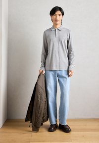 Polo grigio a maniche lunghe, jeans azzurri chiari, giacca trapuntata marrone tenuta in mano e calzature scure. Tessuto morbido con design semplice, dettagli minimi.