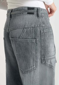 Jean cargo en denim gris avec une finition légèrement délavée, comportant plusieurs poches et une apparence texturée. Détails de patch arrière visibles.