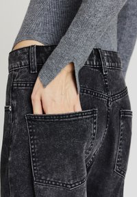 Hand in der Gesäßtasche einer schwarzen, gewaschenen Jeans, gekleidet mit einem grauen, gerippten Langarmoberteil mit einem kleinen Schlitz am Bund.
