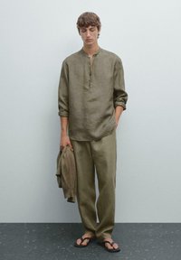 Young man stands against a light gray wall, wearing a loose olive linen shirt, matching pants, and flip-flops, holding a brown sweater.