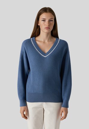 Ung kvinde med langt brunt hår iført en blå V-hals trøje med hvide kanter og off-white bukser, der står mod en ensfarvet grå baggrund.
