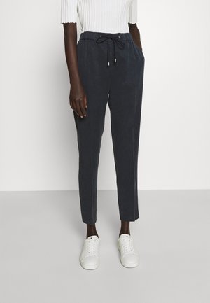 Osoba ubrana w czarne spodnie ze sznurkiem, biały prążkowany top i białe tenisówki, stojąca na tle jednolitego jasnego tła.