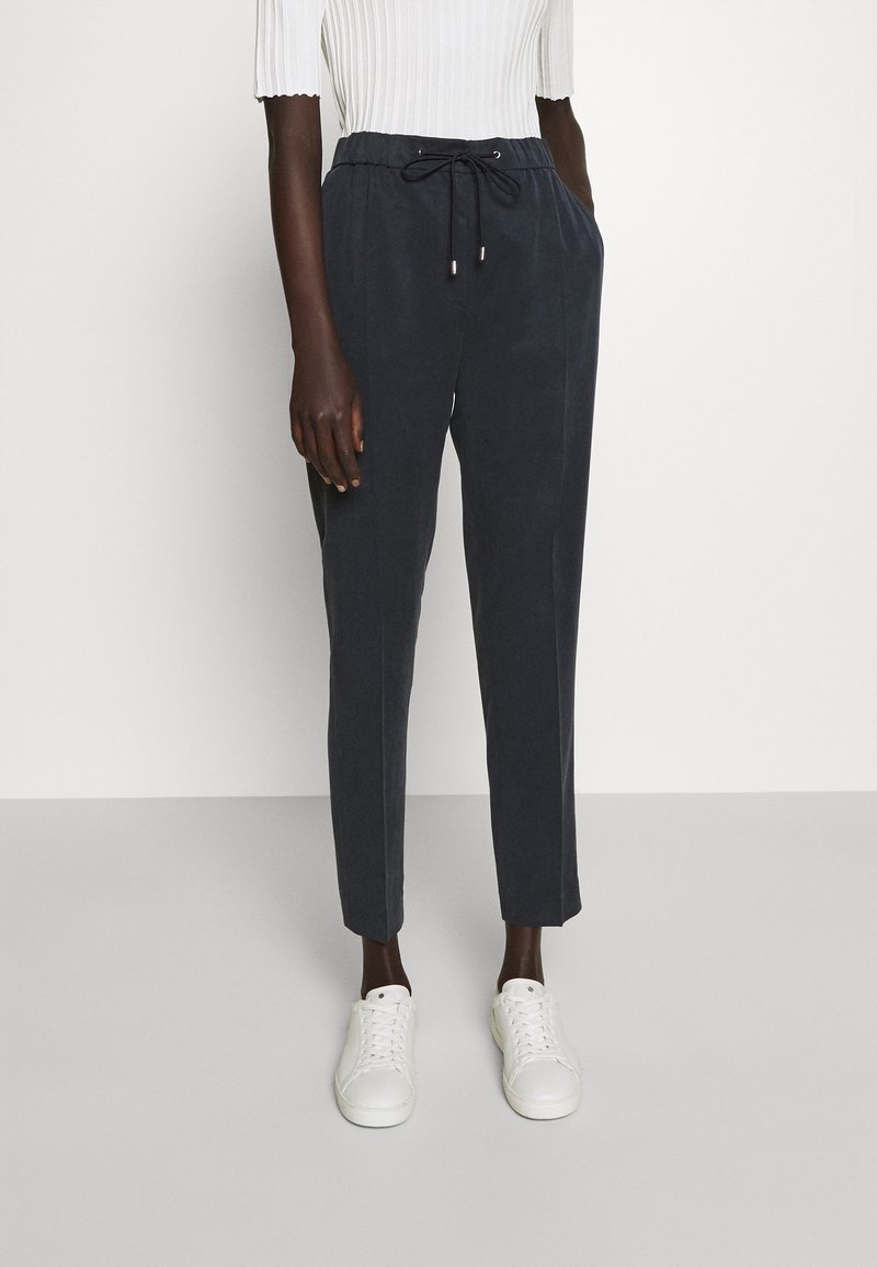 Personne portant un pantalon noir à cordon, un haut blanc côtelé et des baskets blanches, debout devant un fond clair uni.