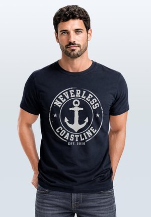 Mann mit dunklen lockigen Haaren und Bart, der ein schwarzes T-Shirt mit einem weißen Anker und den Worten "Nevertheless Coastline Est. 2016" trägt, steht mit den Händen in den Taschen.