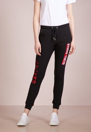 Personne portant un jogging noir avec le texte rouge "STUDIO TOKYO" sur la jambe droite et des caractères japonais sur la jambe gauche, associé à des baskets blanches.