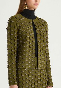Veste en maille verte avec une texture tridimensionnelle en forme d'ondulations. Elle présente un col rond et une ouverture frontale. Associée à une jupe assortie.