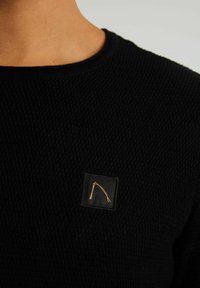 Svart stickad tröja med ett strukturerat mönster och en fyrkantig svart etikett med ett guldgrafiskt designelement på bröstet.