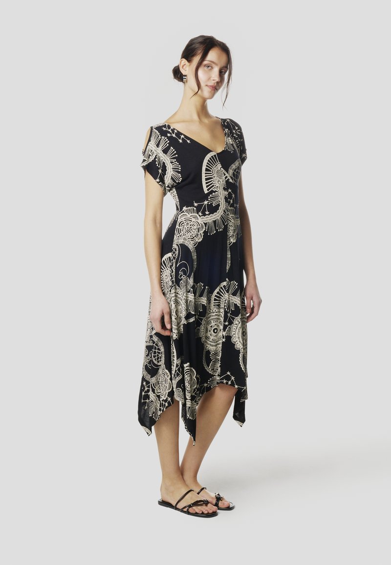 Fekete ruha bonyolult bézs mintákkal, rövid ujjakkal és V-nyakkal. Aszimmetrikus szegély és puha anyag. Fekete lapos szandállal párosítva.