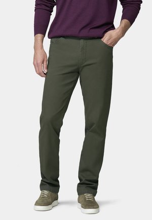 Mann trägt dunkelgrüne Freizeithose, olivgrüne Sneakers und ein lila langärmeliges Hemd, steht mit einer Hand in der Tasche.