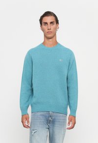 Mann, der einen hellblauen Strickpullover mit kleinem Logo auf der Brust und hellblauen, ausgewaschenen Jeans trägt, steht vor einem schlichten weißen Hintergrund.
