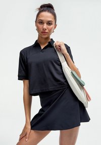 Černá zkrácená polokošile s krátkými rukávy, spárovaná s černou áčkovou sukní. Modelka nese bílou kabelku s barevnými akcenty.