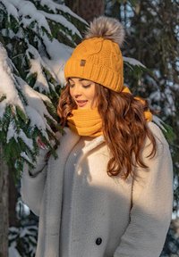 Knit yellow beanie with a gray pom-pom and textured pattern, paired with a matching scarf; model wearing a cream fleece coat. Snowy background.