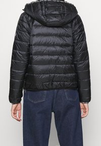 Chaqueta acolchada negra con capucha, que presenta secciones acolchadas horizontales y un detalle de logo en la capucha. Lleva puesta sobre pantalones de mezclilla oscuros.