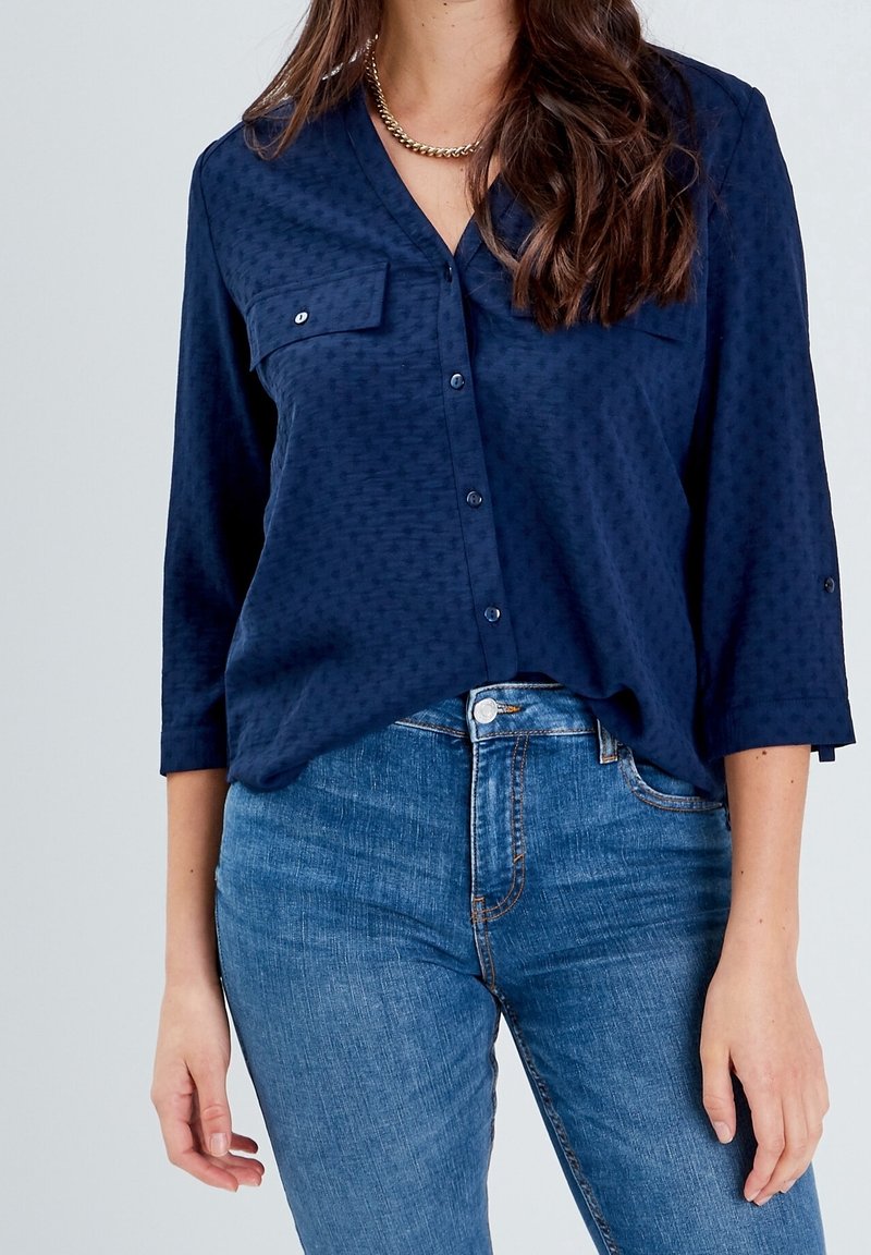 Femme portant une blouse bleu marine à boutons avec des poches poitrine et un jean en denim bleu clair, se tenant contre un fond uni.