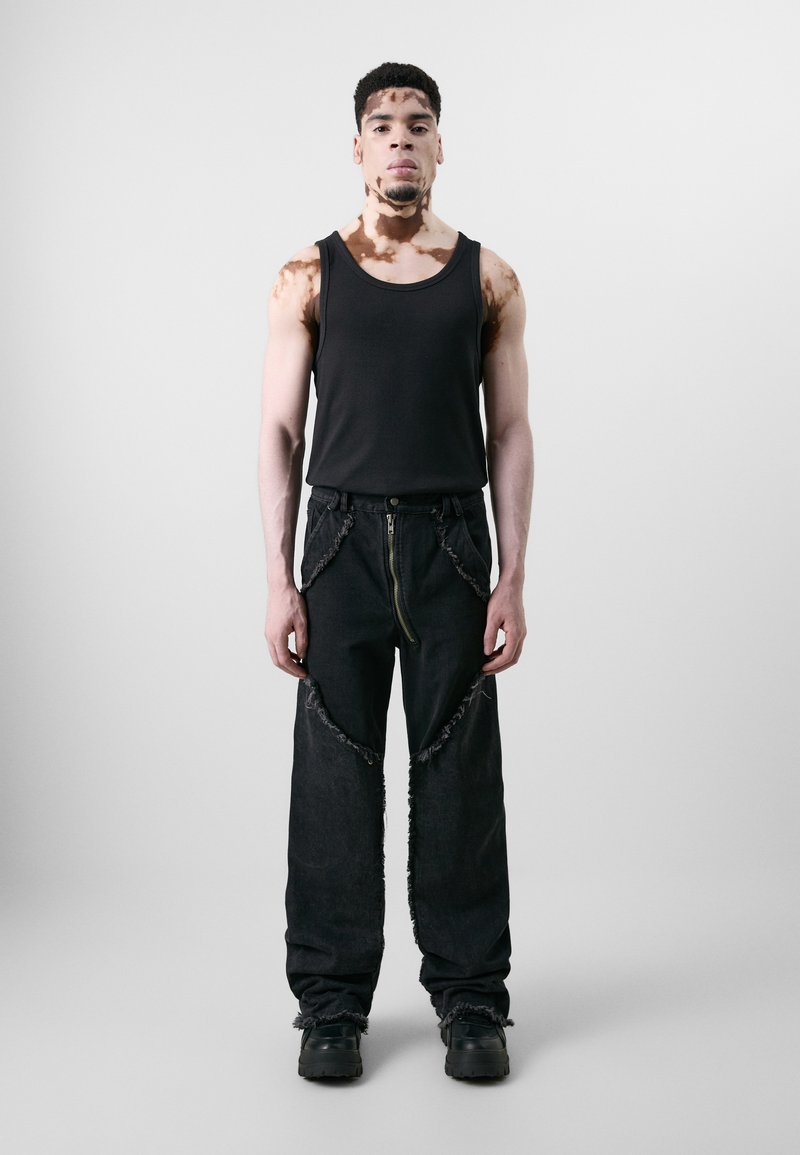 Top noir sans manches et pantalon en denim noir oversize avec des bords effilochés et une fermeture éclair à l'avant. Le mannequin porte des chaussures noires chunky, se tenant contre un fond uni.