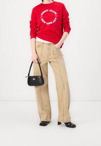 Persona vestida con un sudadera roja de "Tommy Jeans New York City", pantalones anchos beige, zapatos de tacón negros y sosteniendo un pequeño bolso negro.