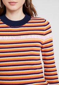 Sweter z prążkowanej dzianiny w wielokolorowe paski, z granatowym dekoltem, w przeważającej różowej, żółtej i czarnej kolorystyce, o dopasowanej sylwetce.