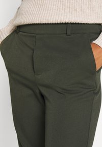 Olivgröna skräddarsydda byxor med en slät yta, platt front, sidofickor och en strukturerad midja, delvis dolda av en lätt stickad tröja.