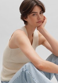 Beige gebreide mouwloze top met een gestructureerd ontwerp. Het model draagt lichtblauwe high-waisted jeans en zit met een neutrale achtergrond.