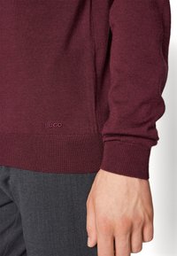 Main reposant sur le côté, portant un pull en maille bordeaux avec le logo "HUGO" près de l'ourlet et un pantalon gris foncé.