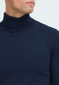 Hombre con un suéter de cuello alto acanalado en azul marino, mostrado desde el cuello hasta la parte superior del pecho, mirando ligeramente de lado contra un fondo liso.