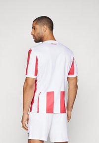 Hvit sportsdrakt med røde vertikale striper, korte ermer og rund hals. Kombinert med hvite shorts.