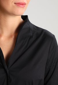 Chemise noire à col, en tissu léger, avec une texture lisse et un plastron avant sans boutons. Apparence nette et ajustée.