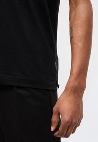 Χέρι και βραχίονας ενός ατόμου που φοράει μαύρο παντελόνι και μαύρο πουκάμισο με μια μικρή ετικέτα «Marc O'Polo» στο τελείωμα, σε απλό φόντο.