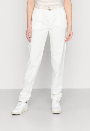 Person iført hvide bukser med lige ben, hvidt flettet bælte, hvide sneakers og en stribet top, der står mod en ensfarvet lys baggrund.