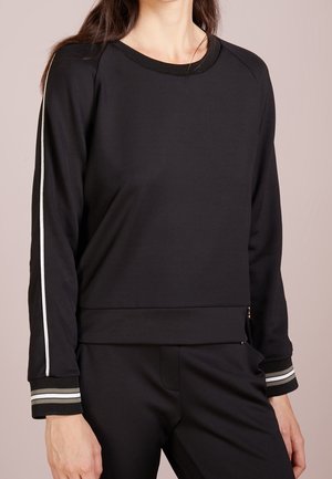 Kobieta ubrana w czarną bluzkę z długim rękawem z białymi paskami na rękawach oraz pasujące czarne spodnie na neutralnym tle.