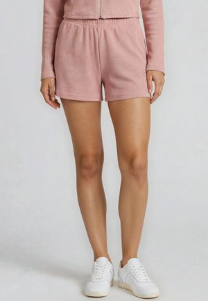 Vrouw draagt zachte roze gestructureerde shorts en bijpassende top met lange mouwen, gecombineerd met witte vetersneakers, staand tegen een effen achtergrond.