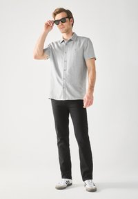 Camisa gris de manga corta con botones, combinada con pantalones vaqueros negros y zapatillas blancas. La camisa presenta una textura sutil y un cuello.