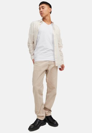 Junger Mann, der ein weißes V-Ausschnitt-Shirt, ein offenes beige gestreiftes Hemd, beigefarbene Hosen und schwarze Schuhe trägt, vor einem schlichten weißen Hintergrund steht.