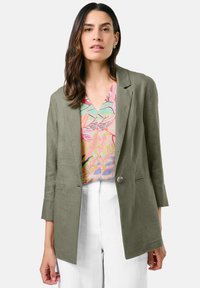 Olivgrüner Leinenblazer mit einem einzigen Knopf, Reverskragen und Seitentaschen, getragen über einem bunten, gemusterten Oberteil und weißen Hosen.
