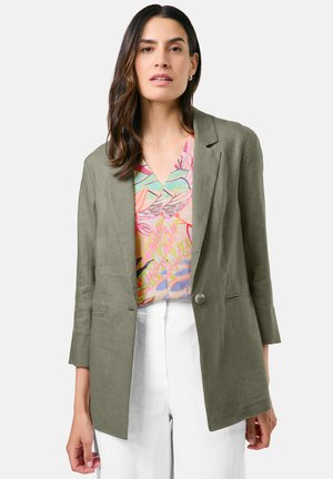 Olivgrüner Leinenblazer mit einem einzigen Knopf, Reverskragen und Seitentaschen, getragen über einem bunten, gemusterten Oberteil und weißen Hosen.