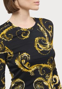Svart langermet skjorte med intrikate gullsurrende mønstre. Rund hals, tettsittende fasong og glatt tekstur.