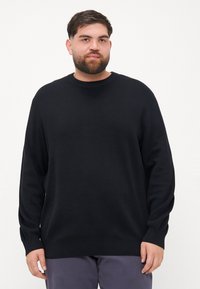 Pulover negru tricotat cu guler rotund, mâneci lungi și manșete cu dungi; textură netedă și croială lejeră. Purtați cu pantaloni gri.