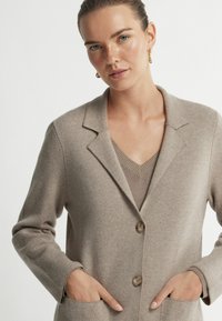 Beżowy sweter z dzianiny z kołnierzykiem, dwoma przednimi kieszeniami i dwoma guzikami. Wewnętrzna warstwa to jasnobrązowy top w serek. Miękka tekstura.