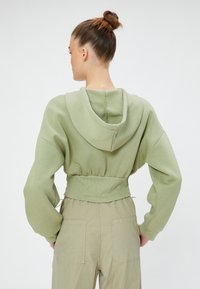 Sudadera verde corta con capucha ajustable, mangas anchas y dobladillo de canalé. Textura suave, combinada con pantalones cargo de color claro.