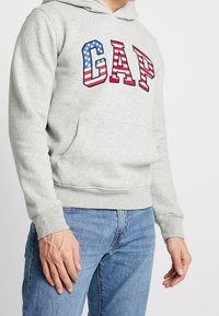 Sudadera con capucha gris que presenta el logo de "GAP" en rojo y azul con un diseño de estrellas y rayas, equipada con un bolsillo frontal y puños de canalé.