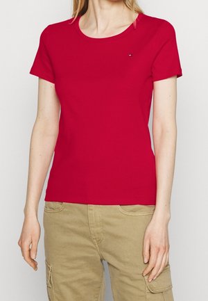 Vrouw draagt een effen rood T-shirt met korte mouwen en beige cargobroek, staat met de armen ontspannen langs het lichaam.