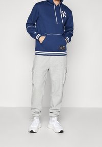 Felpa con cappuccio blu navy con logo bianco, lacci e polsini a strisce abbinata a pantaloni cargo grigi. Scarpe da ginnastica bianche completano l'outfit.