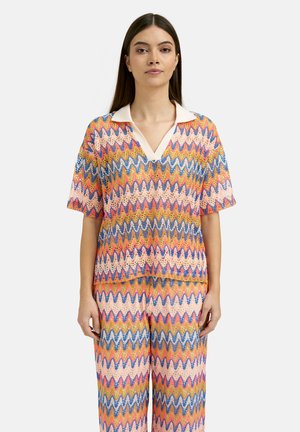 Woman wearing a colorful zigzag patterned short-sleeve shirt with a white collar and matching wide-leg pants, standing against a plain background.