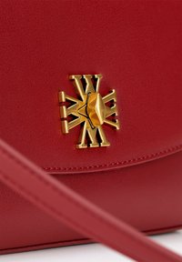 Rode leren handtas met een gestructureerd oppervlak. Heeft een opvallend gouden embleem aan de voorkant, wat detail aan het ontwerp toevoegt.