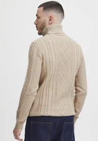 Beige kabelstrikket genser med høy krage, som har teksturerte mønstre og ribbestrikkede mansjetter, synlig søm og en tettsittende design.