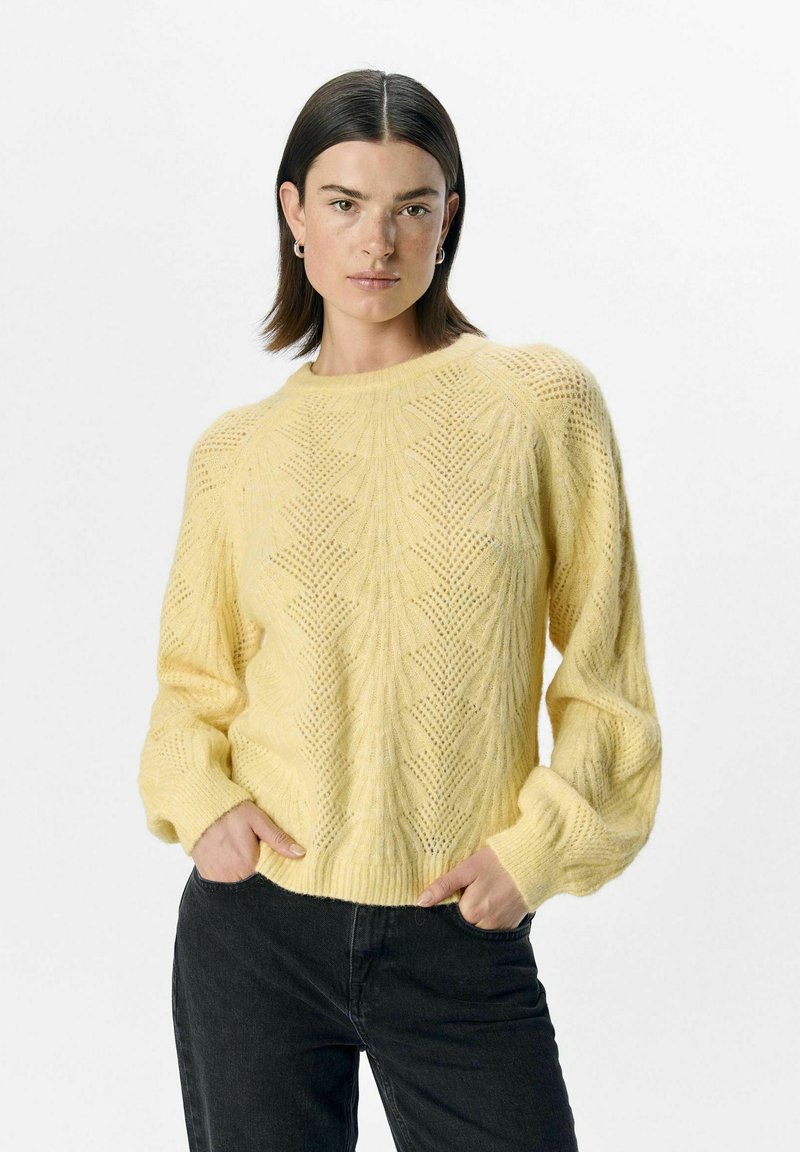 Yellow knitted sweater with a textured pattern, round neckline, and ribbed cuffs. Paired with dark jeans, showcased against a plain background.