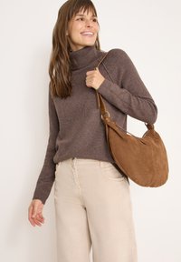 Sac à épaule en suède marron avec une forme courbée et une sangle beige. Le modèle porte un confortable pull à col roulé marron et un pantalon large beige clair.