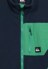 Μπλε φλις μπουφάν σε ναυτικό μπλε με π ράσινο φερμουάρ και τσέπη στο στήθος, με ένα μικρό μαύρο και άσπρο λογότυπο Quiksilver στο πάτο της τσέπης.