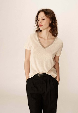 T-shirt beige à col en V avec manches courtes, en matériau texturé, et un col gris contrastant, associé à un pantalon noir taille haute.